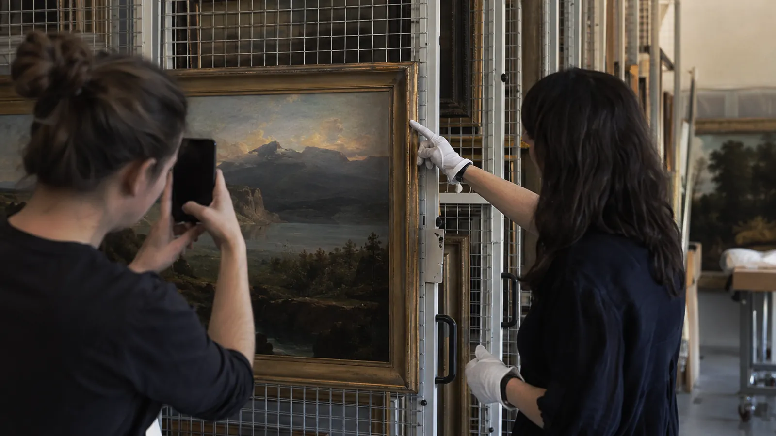 Gloved art handler pointing at the corner of a framed painting while a colleague photographs it